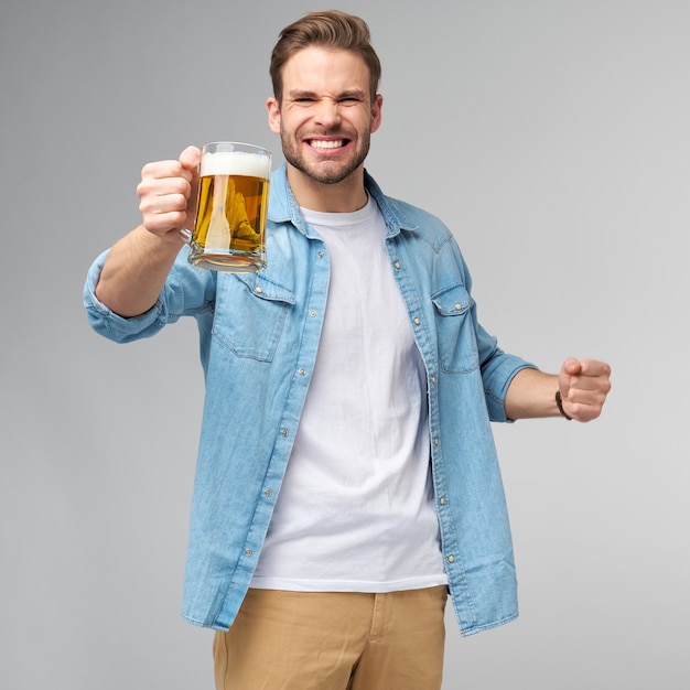 Junger Mann hält tragendes Jeanshemd, das Glas Bier hält, das über graue Wand steht.