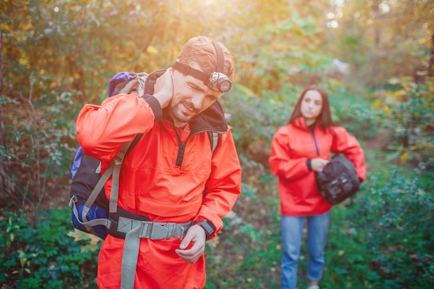 Junger Mann hat Problem in der Halsirea. Er hält die Hand an diesem Ort. Guy leiden. Junge Frau sieht ihn ernst an und hält Schlafsack. Sie tragen orangefarbene Lackets.