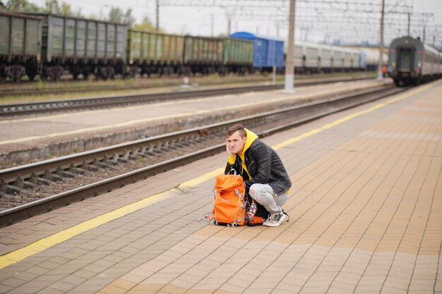 Junger Mann hockt auf dem Bahnsteig und wartet auf den Zug Männlicher Passagier mit Rucksäcken sitzt auf dem Bahnsteig und wartet auf die Zugfahrt Konzept des Tourismus, Reisen und Erholung