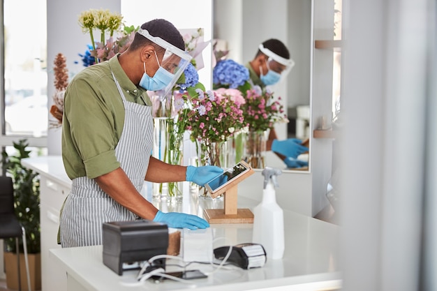 Junger Mann im Antivirenschutz arbeitet an der Theke mit Touchpad und POS-Terminal im Blumenladen