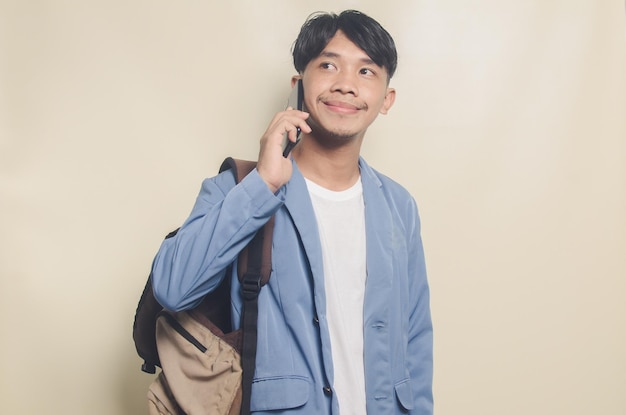 Junger Mann im College-Anzug gestikuliert, einen Freund anrufend, während er einen Rucksack auf isoliertem Hintergrund trägt