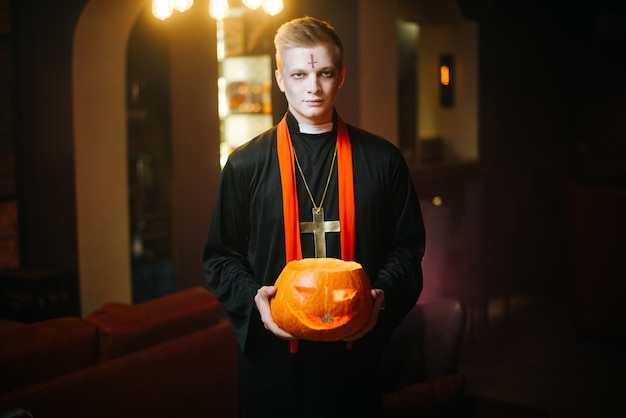 Junger Mann im Halloween-Priesterkostüm, der geschnitzten Kürbis vor der Kamera hält