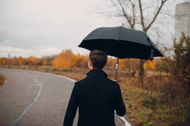 Junger Mann im schwarzen Mantel, der im Regen mit Regenschirm geht