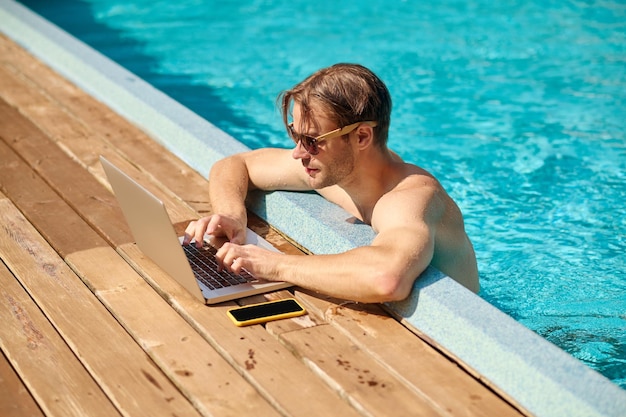 Junger Mann im Schwimmbad, der an einem Laptop arbeitet