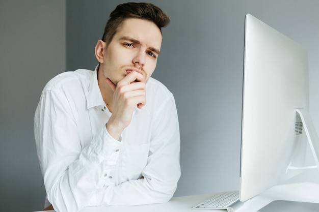 Junger Mann im weißen Hemd, das am Schreibtisch mit einem Computer im Büro arbeitet. Arbeiter auf grauem Hintergrund