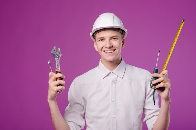 junger Mann im weißen Sturzhelm mit Arbeitsgerät in der Hand auf purpurrotem Hintergrund