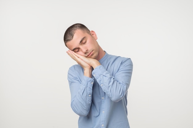 Junger Mann in blauem Outfit, der seine Handflächen wie ein Kissen legt und schläfrig und müde aussieht