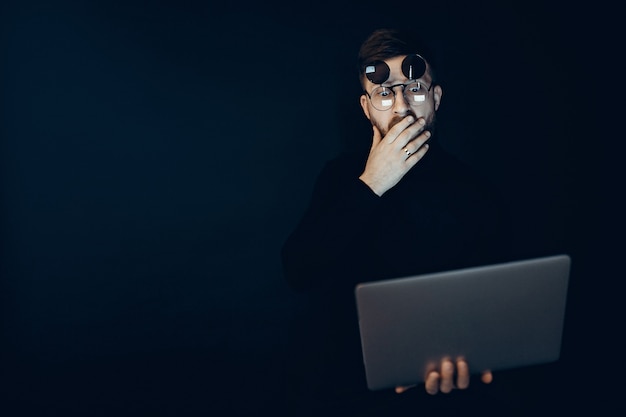 Foto junger mann in den klappgläsern, die den laptop betrachtet es mit schock auf schwarzer wand halten