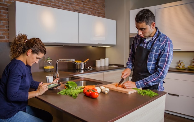 Junger Mann in der heimischen Küche, der Essen zubereitet und Frau sucht Rezept in einem elektronischen Tablet. Modernes Familienlebensstilkonzept.