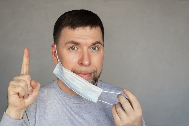 Junger Mann in der medizinischen Maske zeigt mit seinem Zeigefinger auf grauem Hintergrund