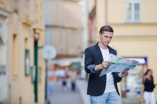 Junger Mann in der Sonnenbrille mit einem Stadtplan und einem Rucksack in Europa. Kaukasischer Tourist, der die Karte der europäischen Stadt auf der Suche nach Anziehungskräften betrachtet.