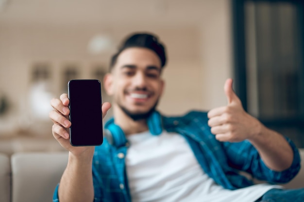 Junger Mann in einem blauen T-Shirt mit einem Telefon in den Händen auf dem Sofa sitzend