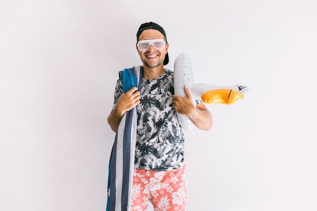 Junger Mann in einem Blumenhemd, einer Mütze, einem Handtuch und einer Brille im Sommer, mit einer Schwimmente, die freudige Gesten macht, bei Tageslicht an einer weißen Wand