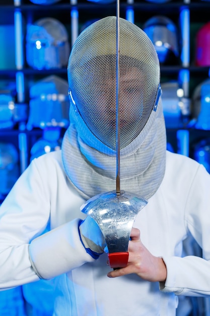 Junger Mann in einem Fechtanzug mit einem Schwert in der Hand, auf blauem Hintergrund, Neonlicht. Der Athlet trainiert. Sport, Jugend, gesunder Lebensstil.