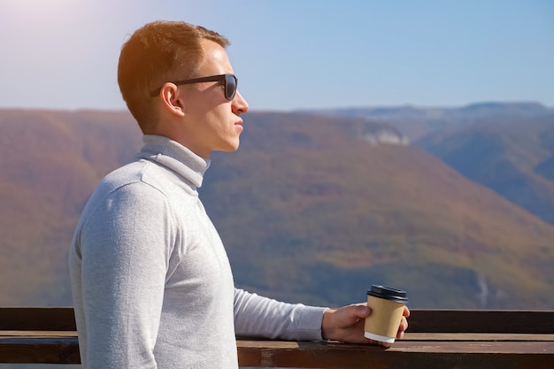Junger Mann in einem grauen Rollkragenpullover und Sonnenbrille trinkt an einem klaren Tag ein Getränk aus einem Plastikglas auf dem Hintergrund der Berge, Sonnenlicht