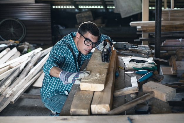 Junger Mann in einem karierten Hemd, Jeans, Schutzbrille und Handschuhen, die manuellen Hobel des Holzes in der Werkstatt planen