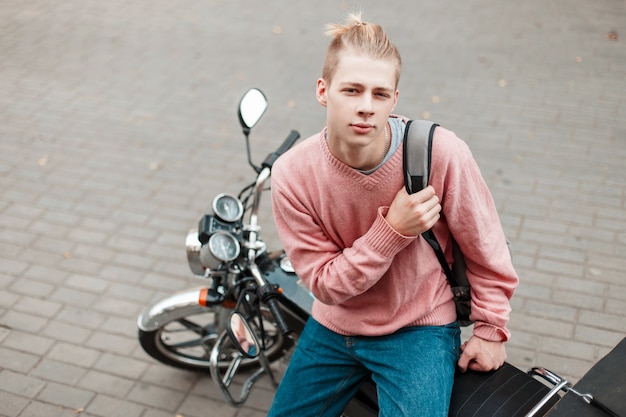 Junger Mann in einem rosa Pullover und einem Rucksack sitzt auf einem Motorrad