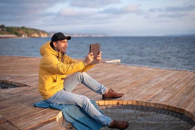 junger Mann in einer gelben Windjacke, schwarze Kappe, die auf einem hölzernen Pier sitzt, macht ein Foto des Sonnenaufgangs auf der Tafel