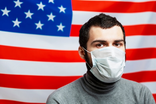 Junger Mann in einer medizinischen Maske auf dem Hintergrund der amerikanischen Flagge. Unabhängigkeitstag, eine Zeit der Unruhe und Pandemie des Coronavirus Nahaufnahme.