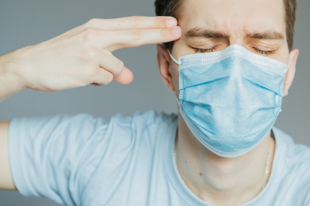 Junger mann in einer medizinischen maske zu hause hält seine finger eine waffe an seiner schläfe. töte, stress