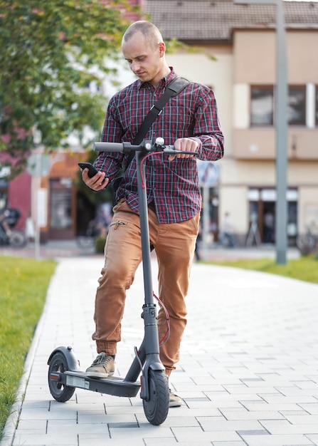 Junger Mann in Freizeithose und Hemd steht neben seinem Elektroroller und checkt sein Handy