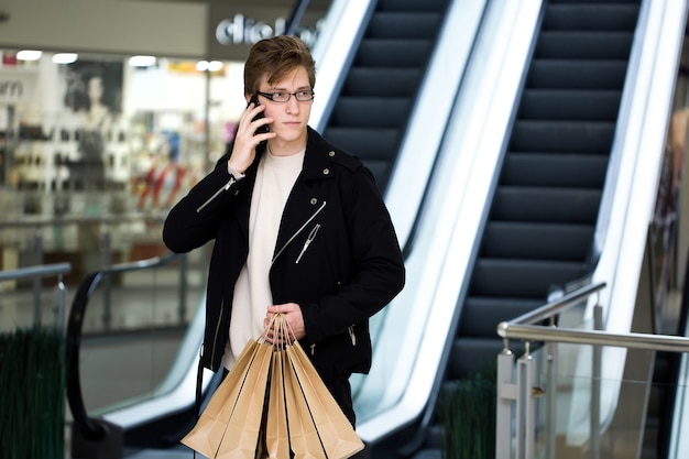 Junger Mann in Gläsern mit Papiertüten, die an der Mall einkaufen und am Telefon sprechen