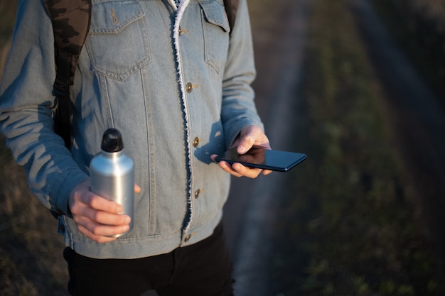 Junger Mann in Jeansjacke mit Smartphone und Aluminiumflasche in seinen Händen