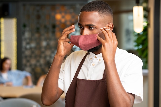 Junger Mann in Kellneruniform, der sich eine Schutzmaske auf das Gesicht legt, während er neue Kunden des Restaurants bedient, in dem er arbeitet