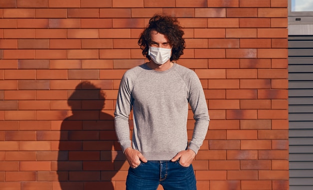 Junger Mann in medizinischer Maske, der die Hände in den Taschen hält und die Kamera anschaut, während er während des Ausbruchs des Coronavirus in der Nähe der Mauer steht
