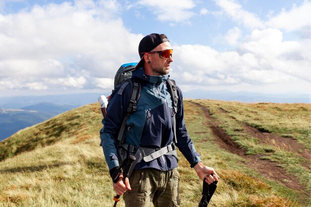 junger Mann in orangefarbener Jacke und Brille geht in den Bergen mit Trekkingstock Touristen-Typ