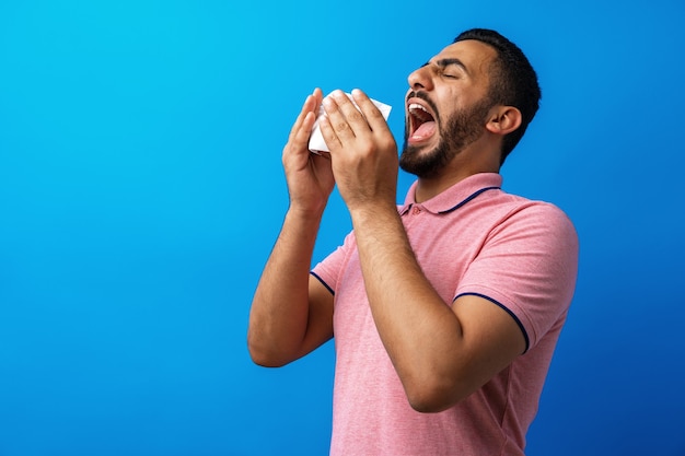 Junger Mann in rosa Hemd mit Allergie oder Erkältung bläst sich die Nase in einem Taschentuch vor blauem Hintergrund