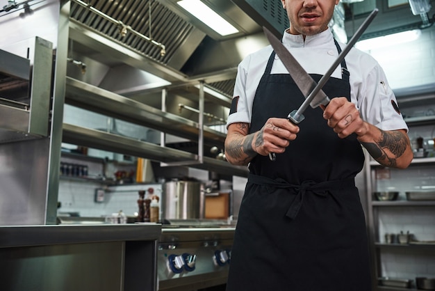 junger Mann in Schürze mit mehreren Tätowierungen auf den Armen, der in einer Restaurantküche ein Messer schärft