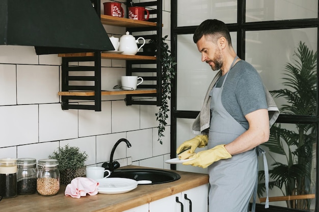 Junger Mann in Schürze und Handschuhen erledigt Hausarbeiten und wäscht Geschirr mit einem Handtuch auf der Schulter. Männer machen Hausarbeit. Haushaltshilfe in stilvoller Küche in moderner Wohnung