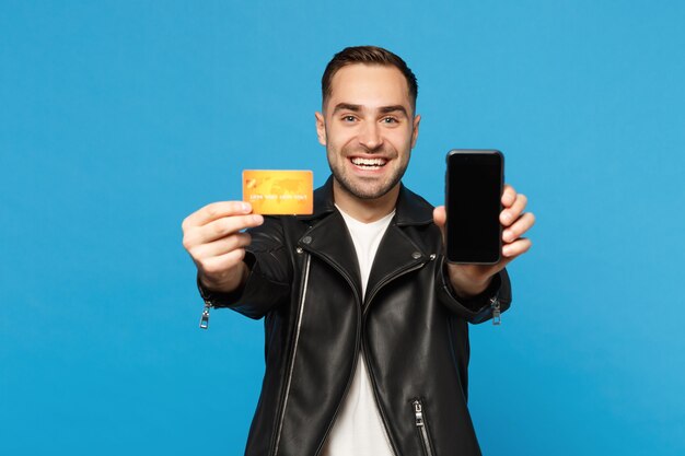 Junger Mann in schwarzer Jacke weißes T-Shirt hält Handy leeren Bildschirm für Werbeinhalte Kreditkarte isoliert auf blauem Wandhintergrund Studioportrait. Menschen-Lifestyle-Konzept Mock-up-Kopienraum