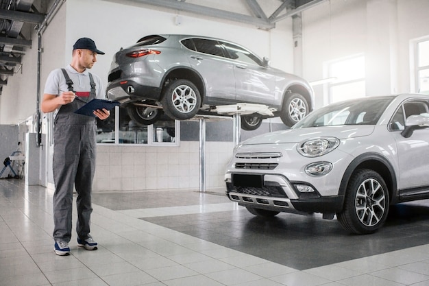 Junger Mann in Uniform steht und schaut Auto an. Er hält Plastiktablette und Stift in den Händen. Woker prüft. Ein anderes Auto steht auf einer Plattform hinter ihm.