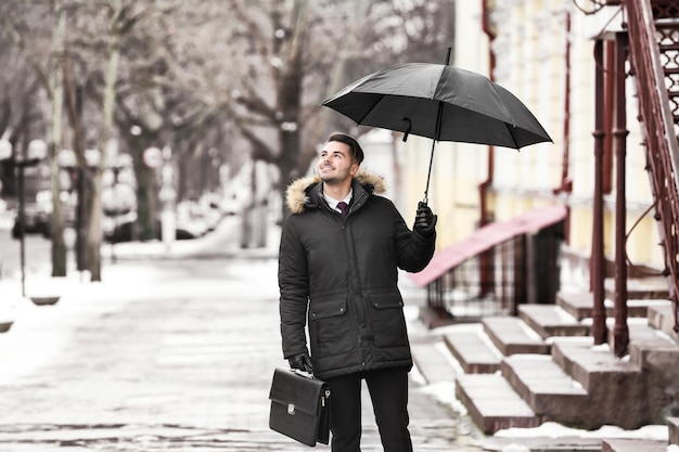 Junger Mann in warmer Kleidung mit dunklem Regenschirm und Aktenkoffer im Freien