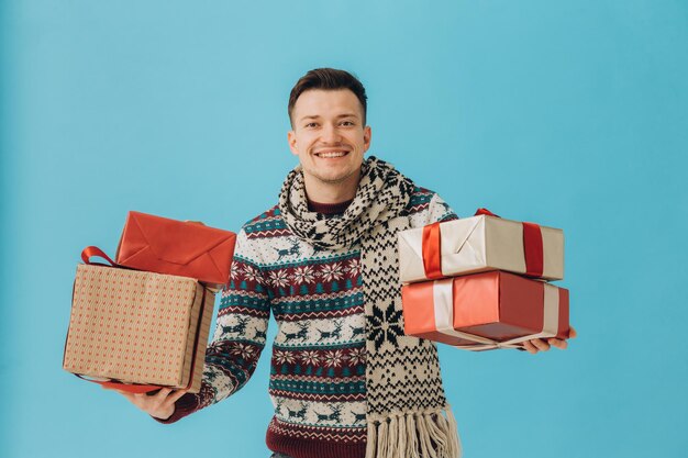 Junger Mann in Weihnachtspullover und -schal, der viele Geschenkboxen mit Geschenkschleife hält, die auf blauem Hintergrund isoliert sind Frohes neues Jahr-Feier-Konzept