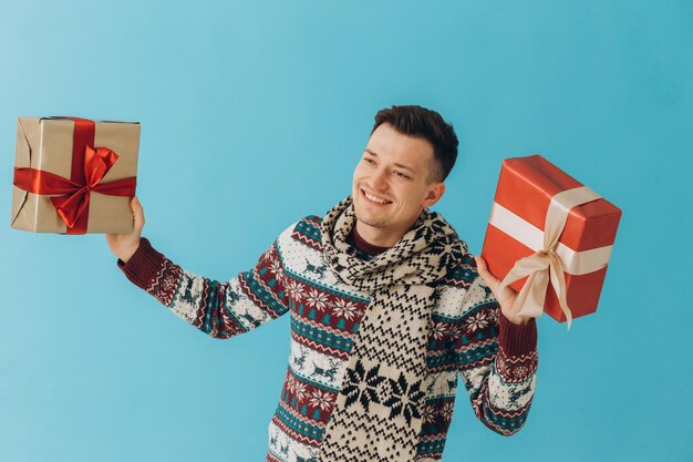 Junger Mann in Weihnachtspullover und -schal, der viele Geschenkboxen mit Geschenkschleife hält, die auf blauem Hintergrund isoliert sind Frohes neues Jahr-Feier-Konzept