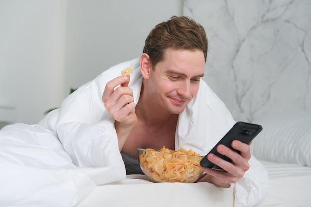 Junger Mann isst Chips im Schlafzimmer mit Fast-Junk-Food