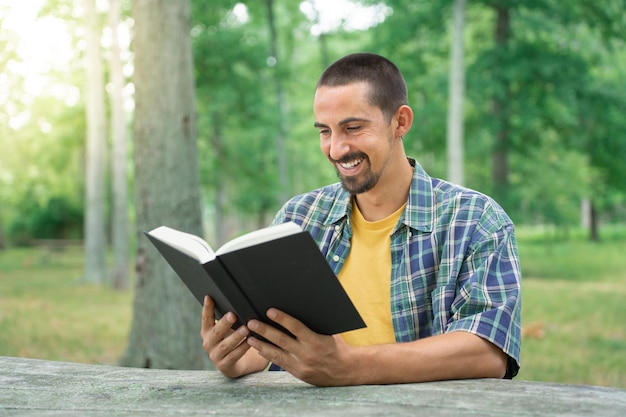 Junger Mann lächelt und liest Buch im grünen Park im Freien. Hobby und Lebensstil. Glaube, Spiritualität.