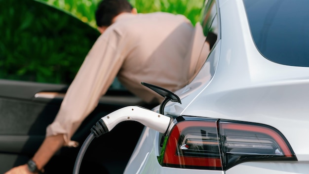 Junger Mann lädt im Frühling im grünen Stadtpark die Batterie eines Elektroautos an der Ladestation auf. Wiederaufladbares Elektroauto für einen nachhaltigen, umweltfreundlichen urbanen Reiselebensstil. Zweckmäßig