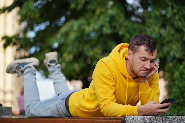 Junger Mann liegt auf dem Bauch draußen auf einer Bank mit hochgelegten Beinen und surft auf ihrem Handy Mann nutzt mit einem Smartphone das Internet im Innenhof eines Wohnhauses