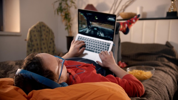 Junger Mann liegt auf dem Bett und spielt Rennvideospiel auf dem Laptop