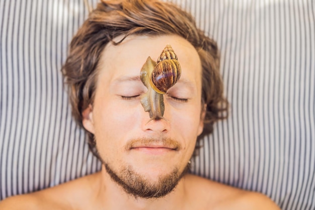 Junger Mann macht eine Gesichtsmaske mit Schneckenschleim Schnecke kriecht auf eine Gesichtsmaske SPA für den Menschen SPA für alle