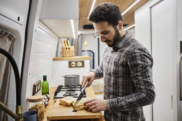 Junger Mann macht Frühstück in winziger Küche am Wohnwagen und schneidet Sandwiches