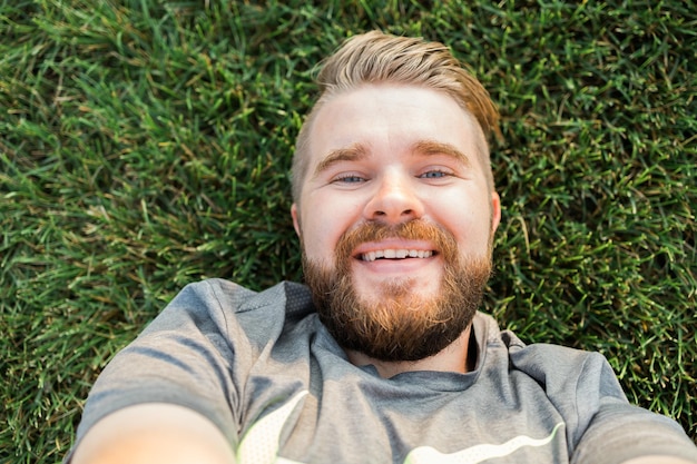 Junger Mann mit Bart und modischer Frisur, der auf einem Gras liegt und ein Selfie mit Smartphone oder Tablet macht und in die Kamera schaut