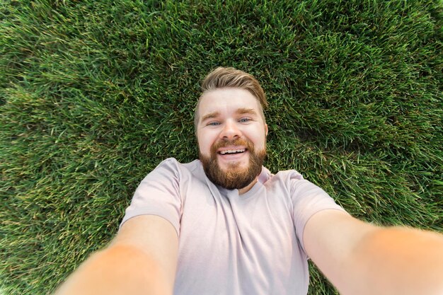 Junger Mann mit Bart und modischer Frisur, der auf Gras liegt und ein Selfie mit Smartphone macht oder