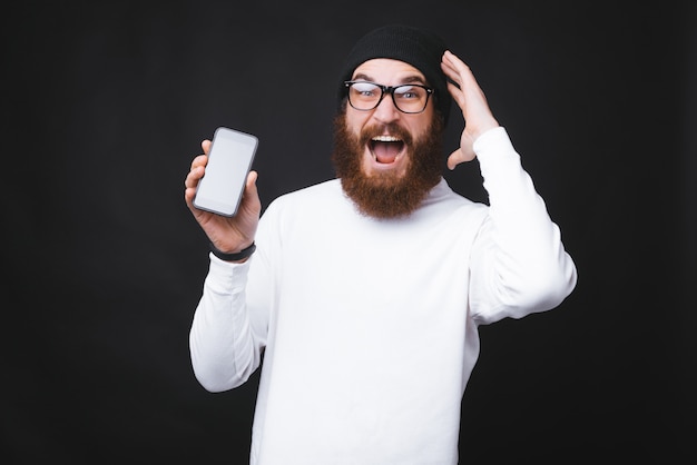 Junger Mann mit Bart wird verrückt und hält ein Telefon in der Nähe der schwarzen Wand.