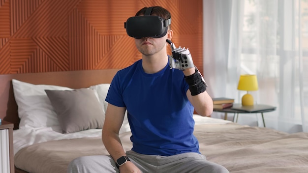 Junger Mann mit bionischer Armprothese und Virtual-Reality-Brille sitzt auf dem Bett