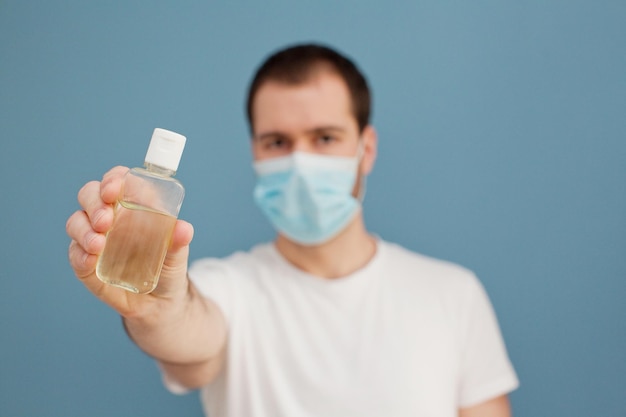 Junger Mann mit chirurgischer Maske verwendet Händedesinfektionsmittel, um seine Hände von Keimen auf blauem Hintergrund zu reinigen